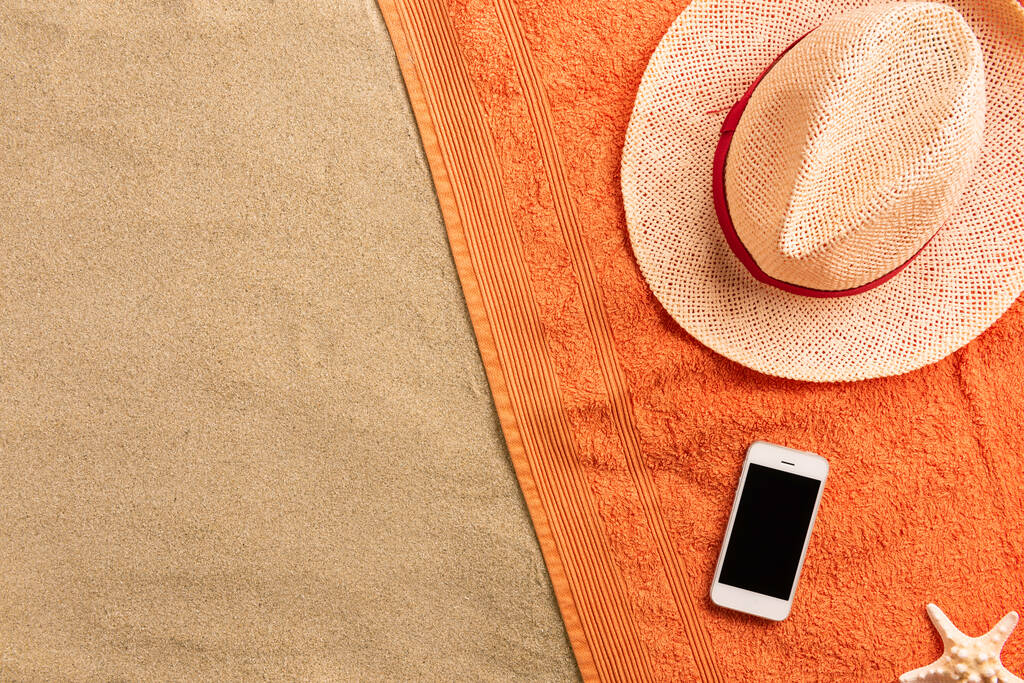 Κορυφαία προβολή ψάθινο καπέλο και smartphone με χώρο αντιγραφής. Αξεσουάρ ταξιδιωτών στην άμμο. Ταξιδιωτικές διακοπές. Θερινό φόντο. Σύνθεση περιγράμματος από πετσέτα - Φωτογραφία, εικόνα