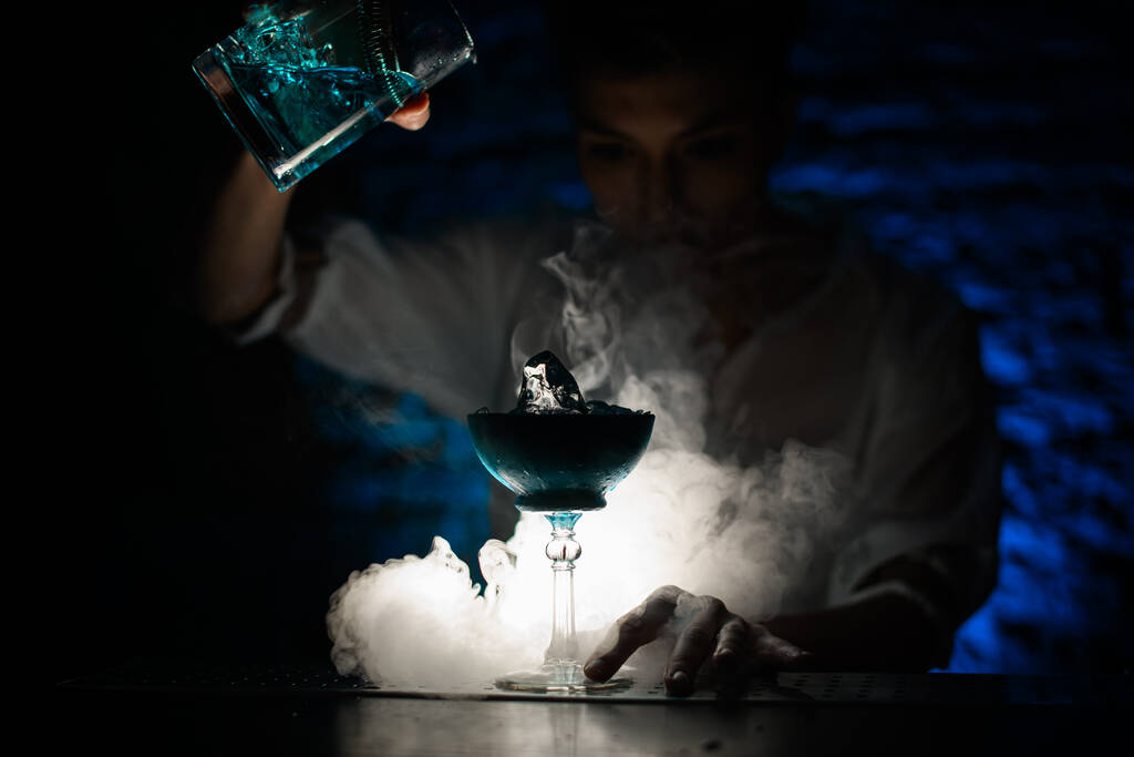 barman nalévá modrý koktejl z míchacího šálku do skla v tmavém kouřovém baru. - Fotografie, Obrázek