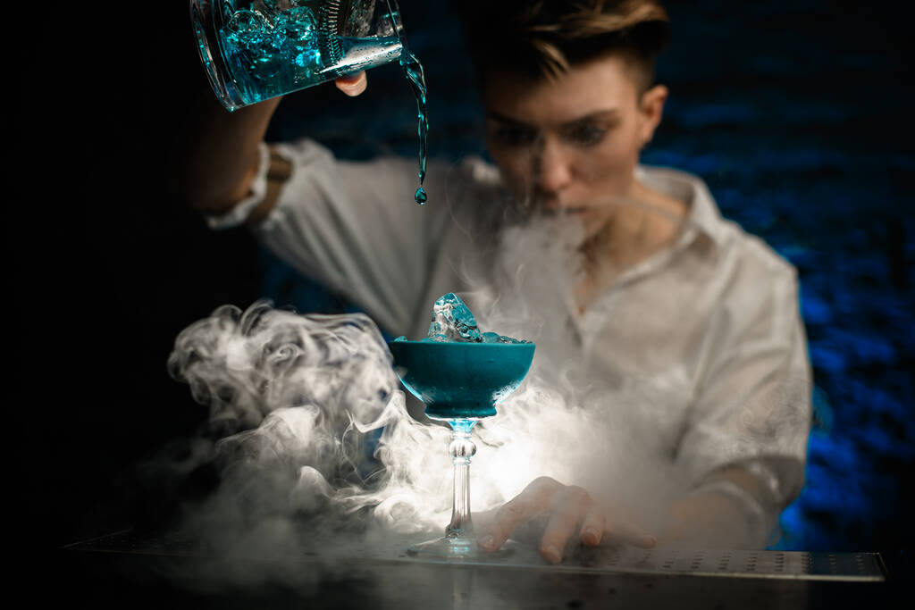 Lähemmäs. Nainen baarimikko kaataa sinisen cocktailin sekoittamalla kupin lasiin savuisessa baarissa ja katso sitä
 - Valokuva, kuva