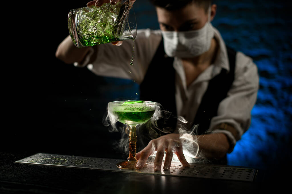 Kobieta barman w masce starannie nalewa zielony koktajl z kubka mieszania do dymu szklanego - Zdjęcie, obraz