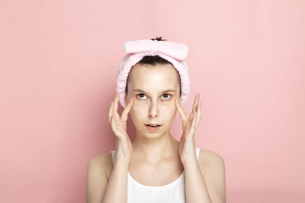 Chica joven con parches en los ojos mira hacia arriba y toca las manos a la cara en el fondo rosa
 - Foto, imagen