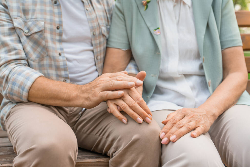 Τραβηγμένη φωτογραφία ηλικιωμένου ζευγαριού να κρατιέται χέρι-χέρι ενώ κάθεται μαζί στο παγκάκι. Εστίαση στα χέρια. - Φωτογραφία, εικόνα