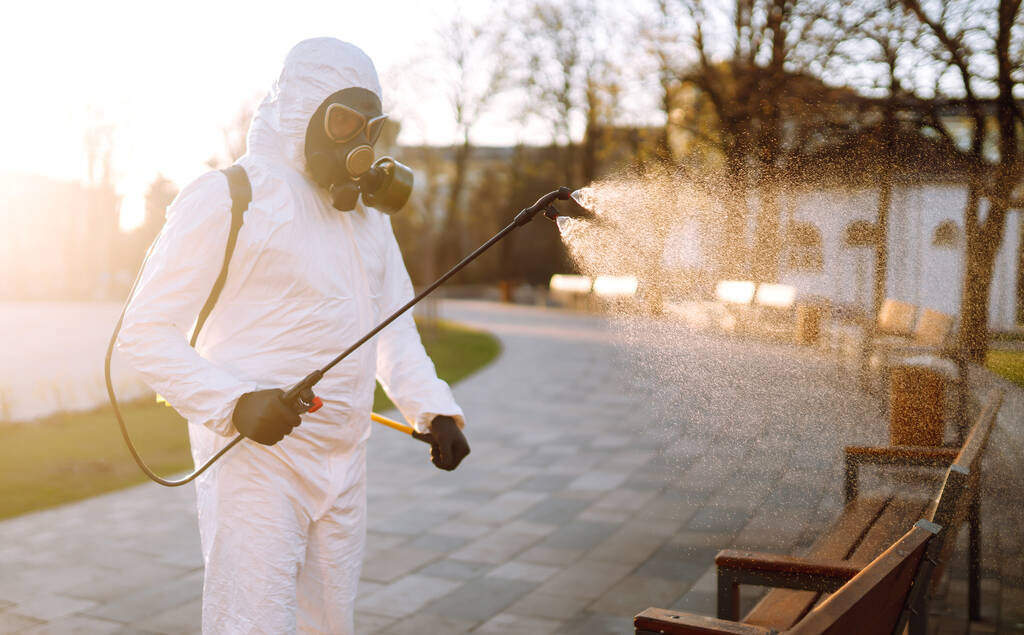 Людина в захисному костюмі дезінфікує громадські місця на сонці хімічними речовинами, щоб запобігти поширенню коронавірусу, пандемії в карантинному місті. Коїд -19. Очищення. - Фото, зображення