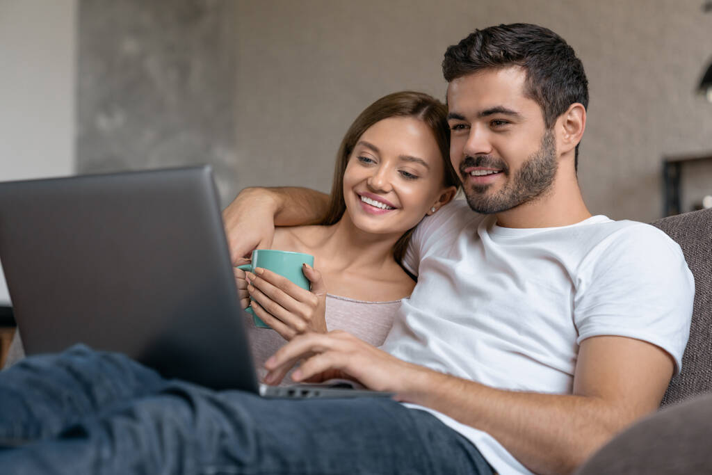 笑顔の若いカップルは、ノートパソコンを使用している間、ソファでリラックスして自宅でお茶を飲む - 写真・画像