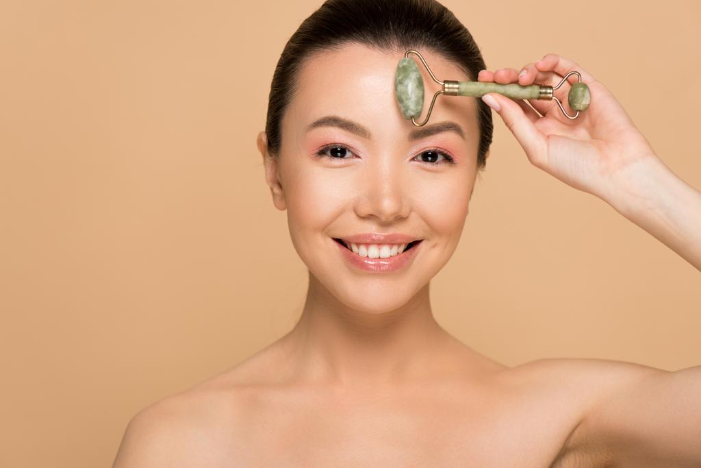 schön nackt asiatische Frau Herstellung Massage des Gesichts mit Jade-Rolle isoliert auf beige - Foto, Bild