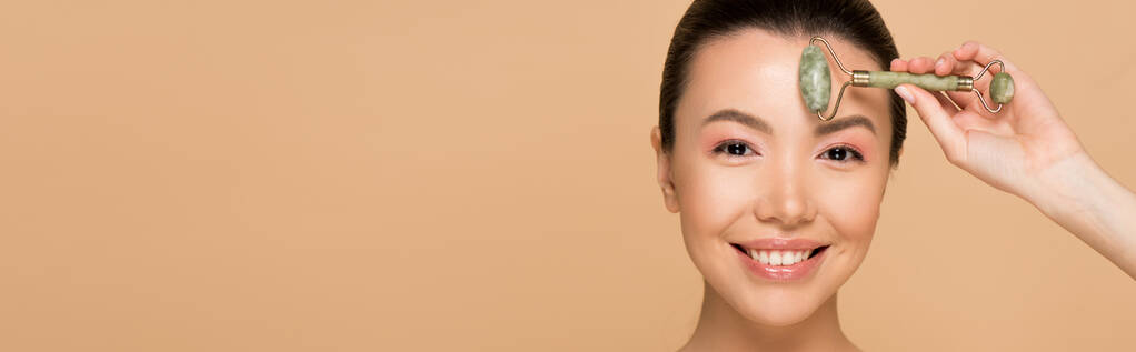 Güzel, gülümseyen Asyalı bir kızın bej rengi bir tekerle yüz masajı yaptığı panoramik bir fotoğraf. - Fotoğraf, Görsel