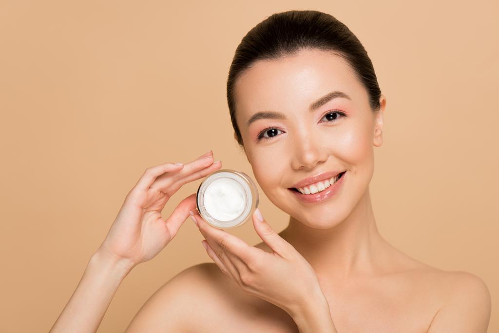 smiling naked asian woman holding glass container with cosmetic cream isolated on beige - Photo, Image