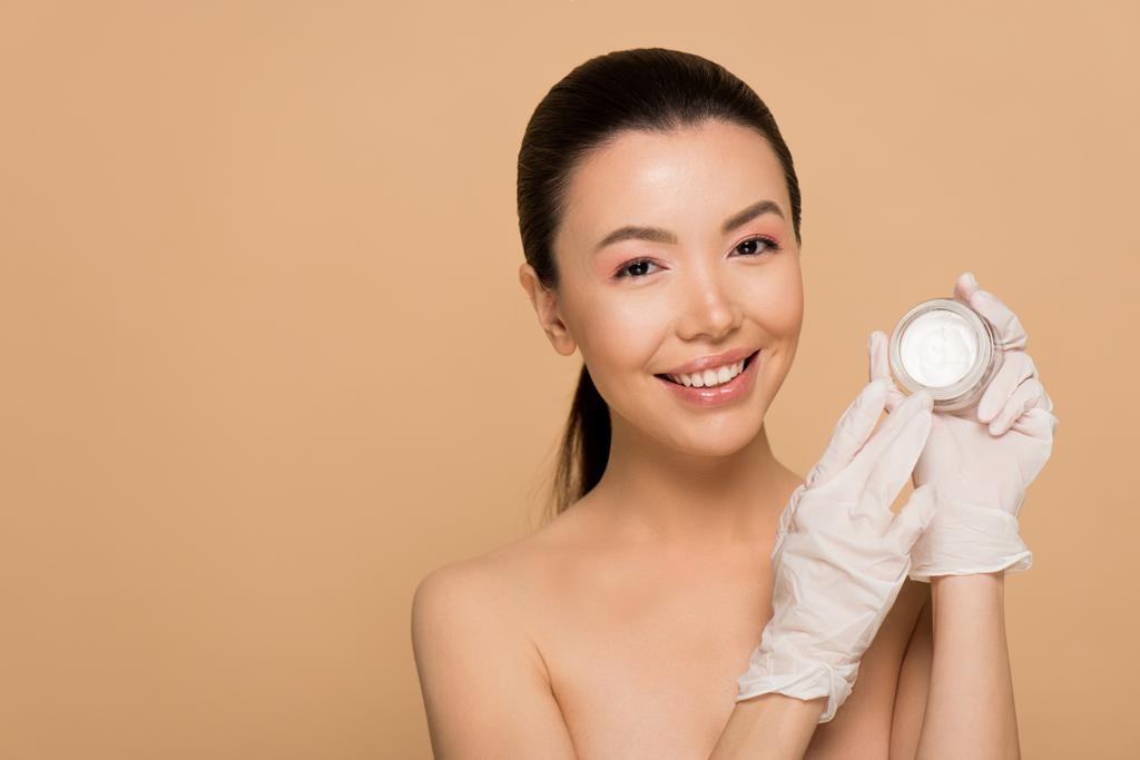 attraktive glückliche nackte asiatische Mädchen in Latex-Handschuhen halten Glascontainer mit Gesichtscreme isoliert auf beige - Foto, Bild