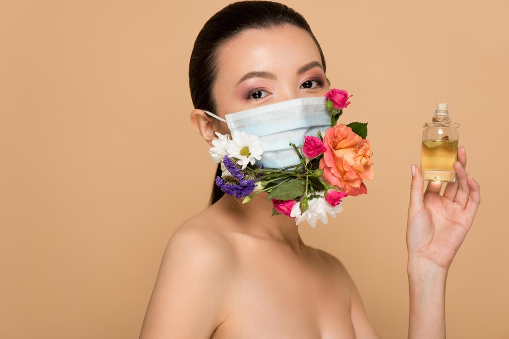 приваблива оголена азіатська дівчина в квітковій масці для обличчя, що тримає скляну пляшку парфумів ізольовано на бежевому
 - Фото, зображення