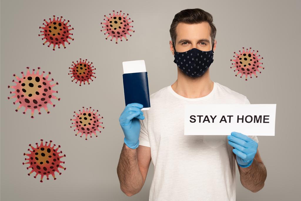 Man in safety mask and latex gloves holding card with stay at home lettering and passport with air ticket isolated on grey, bacteria illustration  - Photo, Image