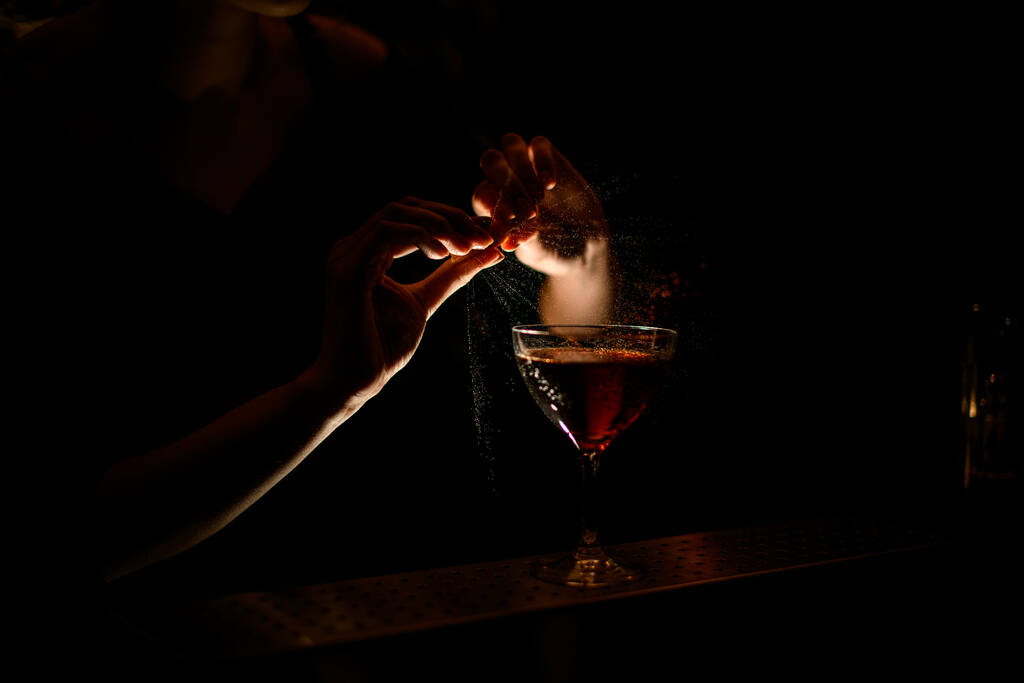 Mulher barman polvilha suco de cítricos em belo copo com vinho no bar escuro
. - Foto, Imagem