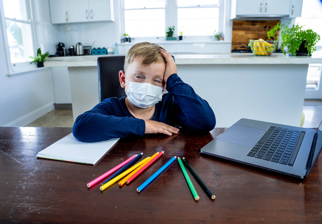 Coronavirus járvány. Lezárás és iskolabezárás. Iskolás fiú arcmaszkkal nézi az online oktatást, unatkozik és depressziós otthon. COVID-19 világjárvány kényszeríti a gyerekeket az online tanulásra. - Fotó, kép