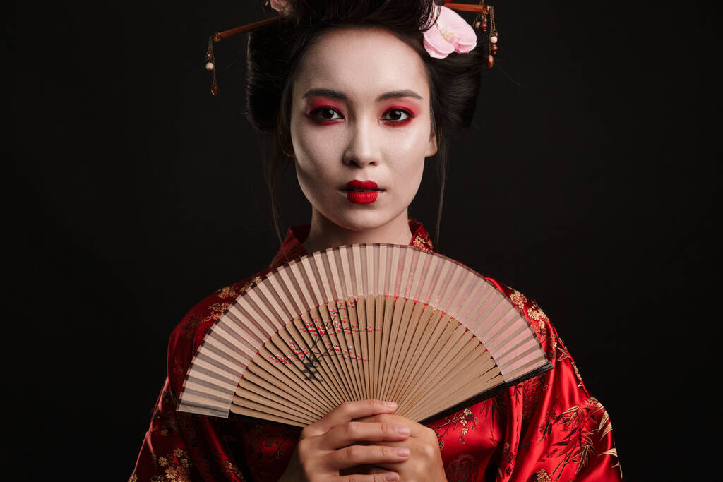 Bild einer jungen Geisha-Frau im traditionellen japanischen Kimono, die einen hölzernen Handfächer über schwarzem Hintergrund hält - Foto, Bild