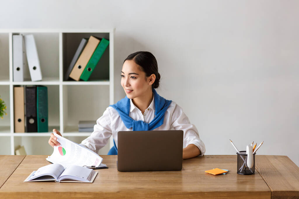 Kép fiatal ázsiai titkárnő ül az asztalnál, és használja laptop számítógép, miközben dolgozik az irodában - Fotó, kép