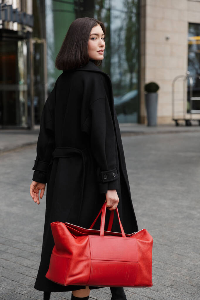 Atraktivní mladá brunetka žena v podzimním kabátě procházky v ulici, nesoucí cestovní tašku - Fotografie, Obrázek