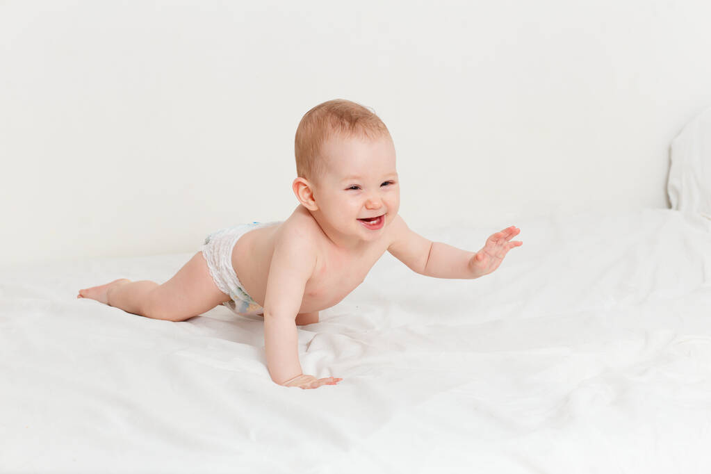 Ένα λευκό μωρό οκτώ μηνών, που γελάει σε μια πάνα, σέρνεται στο κρεβάτι. Ευτυχισμένο γυμνό μωρό. ανάπτυξη των παιδιών έως ένα έτος - Φωτογραφία, εικόνα