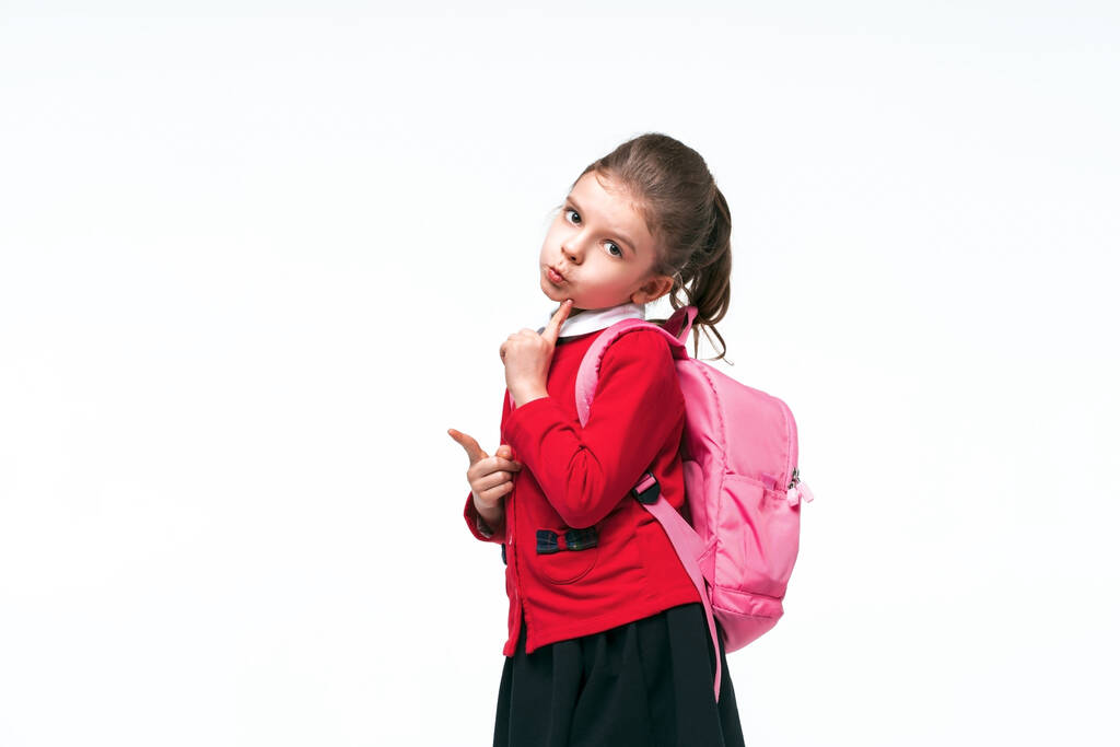 Urocza dziewczynka w czerwonej kurtce szkolnej, czarnej sukience, plecaku wskazującym empy przestrzeni podczas pozowania na białym tle studio. Izolacja - Zdjęcie, obraz