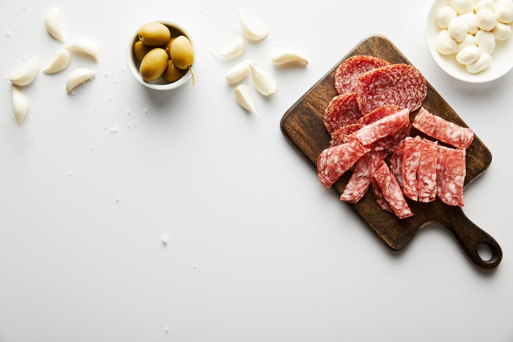 Top view of meat platter, garlic, sea salt and bowls with olives and mozzarella on white background - Photo, Image