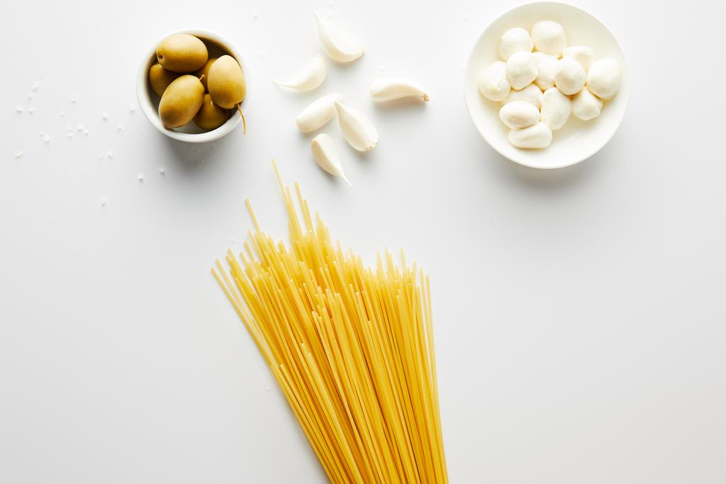 Horní pohled na špagety, česnek, mořskou sůl a misky s olivami a mozzarella na bílém pozadí  - Fotografie, Obrázek