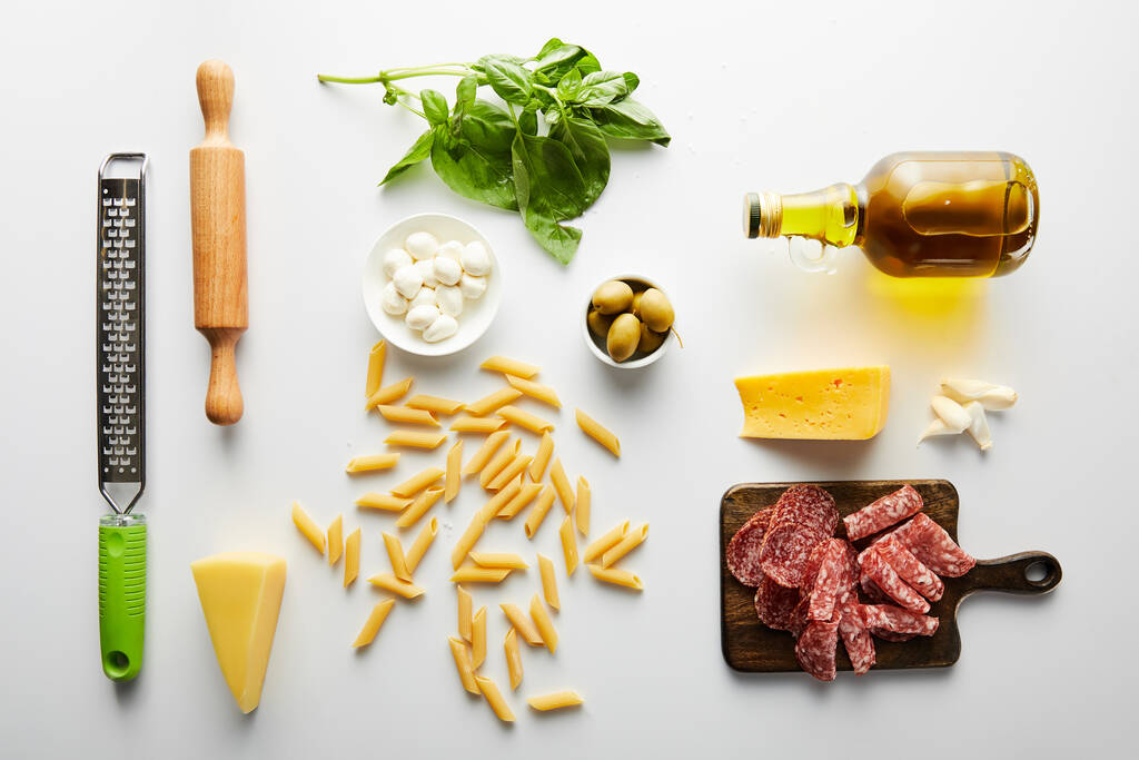Acostado plano con plato de carne, botella de aceite de oliva, rodillo, rallador, pasta e ingredientes en blanco
  - Foto, imagen