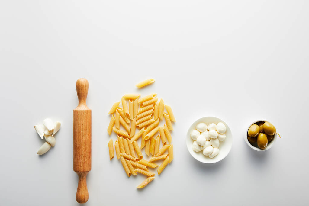 Flat lay with garlic, rolling pin, pasta and bowls with olives and mozzarella on white background  - Photo, Image