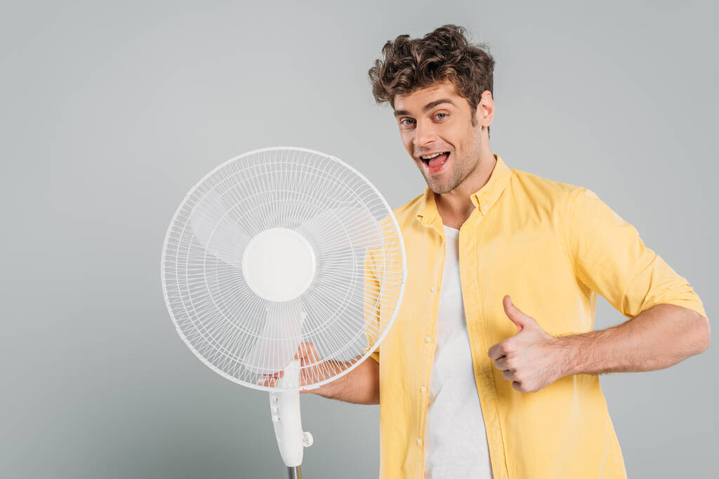 Hombre con la boca abierta sosteniendo ventilador eléctrico, mirando a la cámara y mostrando como signo aislado en gris
 - Foto, imagen