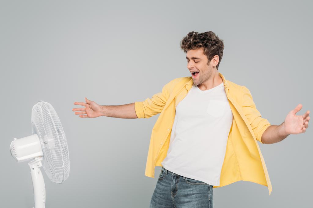 Izgalmas férfi tárt karokkal élvezi elektromos ventilátor elszigetelt szürke - Fotó, kép