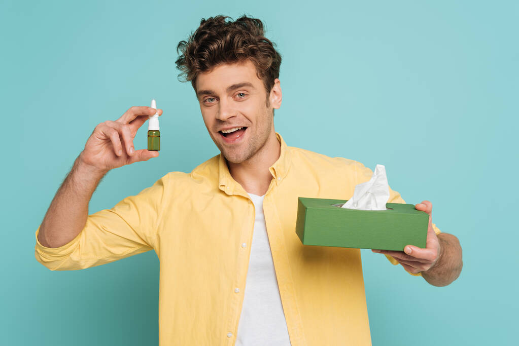 Přední pohled muže usmívá a ukazuje láhev s nosní kapky a box s ubrousky izolované na modré - Fotografie, Obrázek