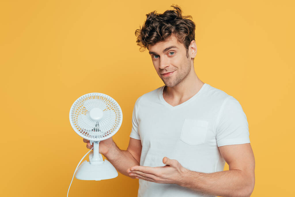 Man smiling, looking at camera and pointing at desk fan isolated on yellow - Photo, Image
