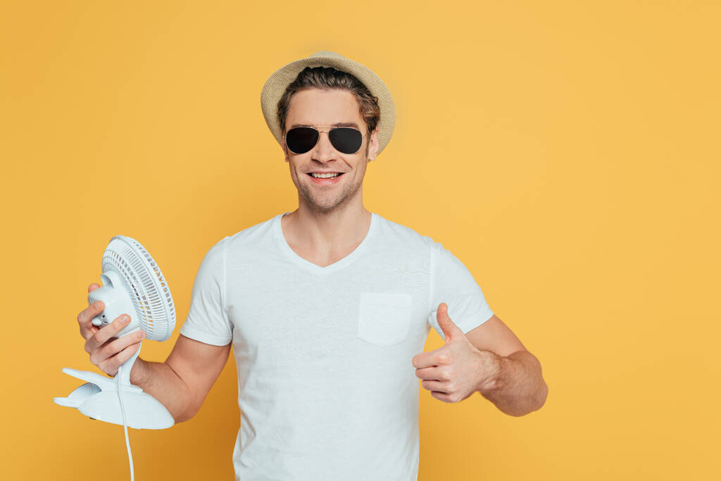 Přední pohled na muže v klobouku a slunečních brýlích s ventilátorem a ukazující jako znamení izolované na žluté - Fotografie, Obrázek