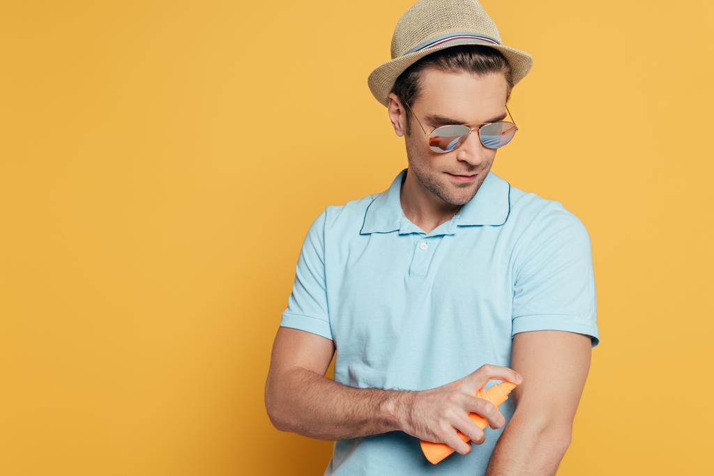 Muž v klobouku a slunečních brýlích nanáší opalovací krém izolovaný na žlutou  - Fotografie, Obrázek