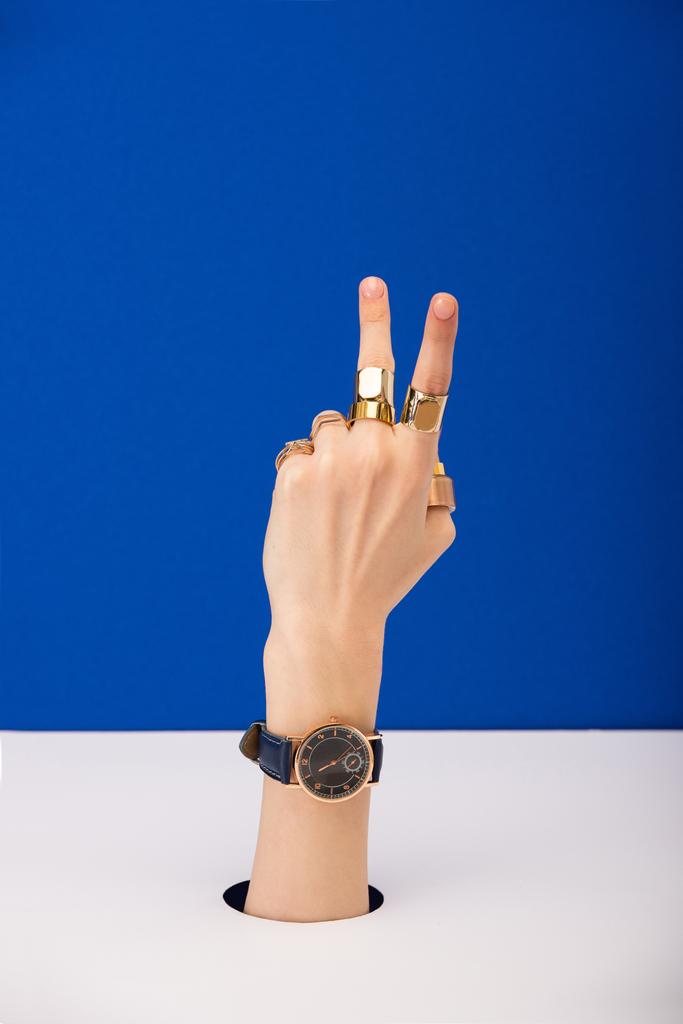 обрезанный вид женщины с наручными часами и кольцами показывая знак мира изолированы на синий
 - Фото, изображение