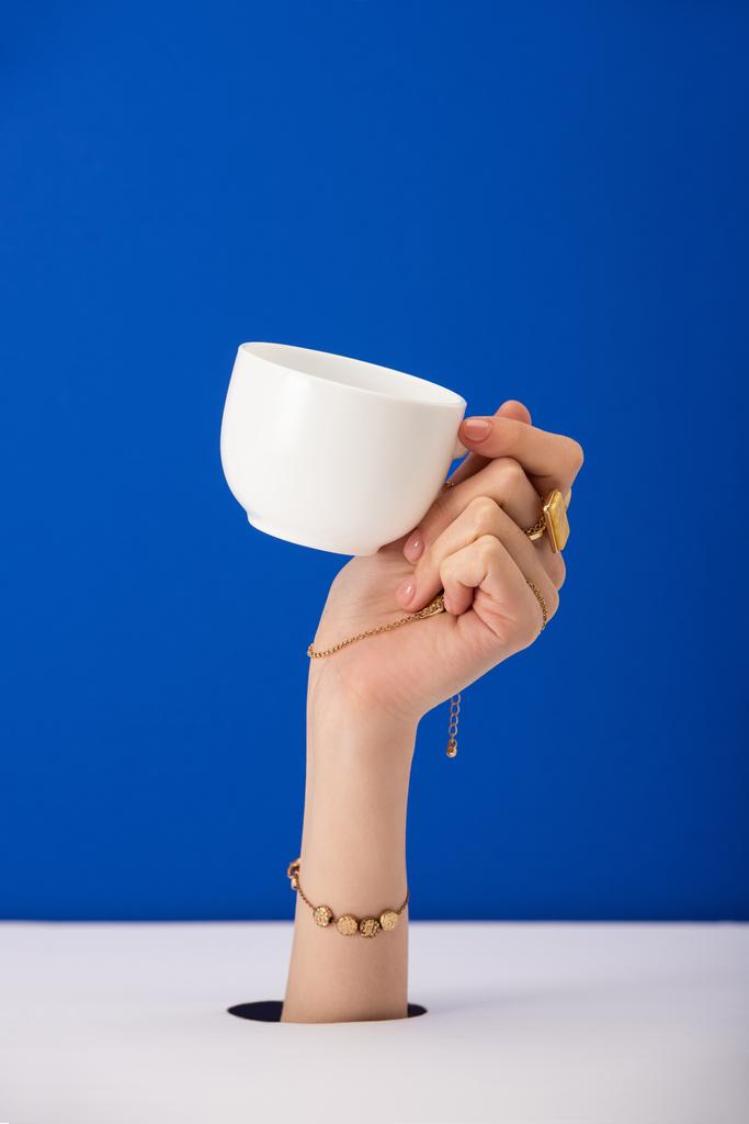 abgeschnittene Ansicht einer Frau mit Armband an der Hand, die eine Tasse Kaffee isoliert auf blau hält - Foto, Bild