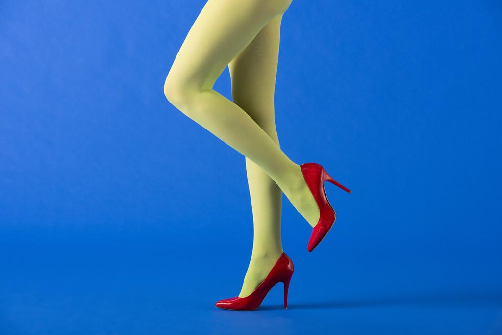 oříznutý pohled na model v zelených punčocháčích a červených podpatcích pózujících na modré  - Fotografie, Obrázek