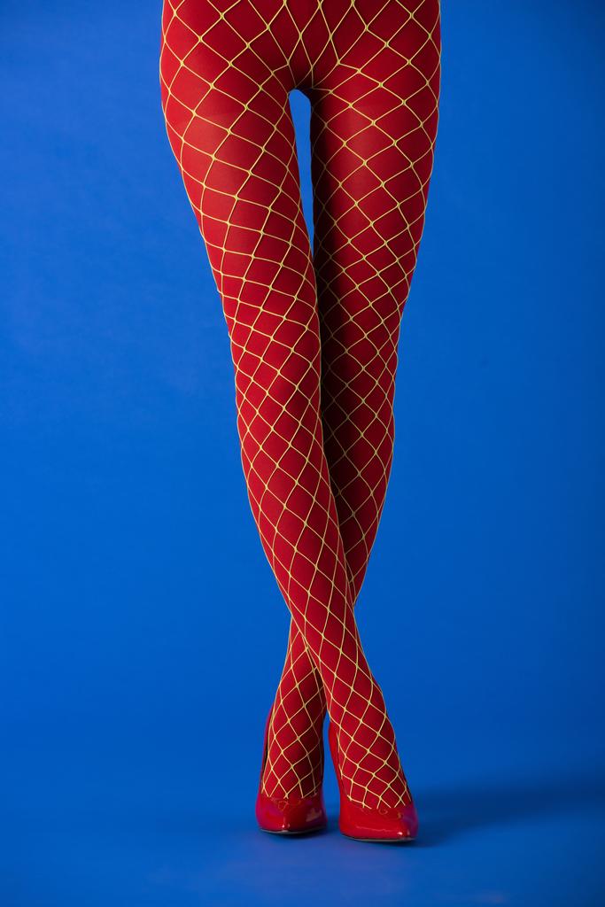 cropped άποψη του μοντέλου σε καλσόν fishnet και κόκκινα τακούνια στέκεται με σταυρωμένα πόδια σε μπλε  - Φωτογραφία, εικόνα