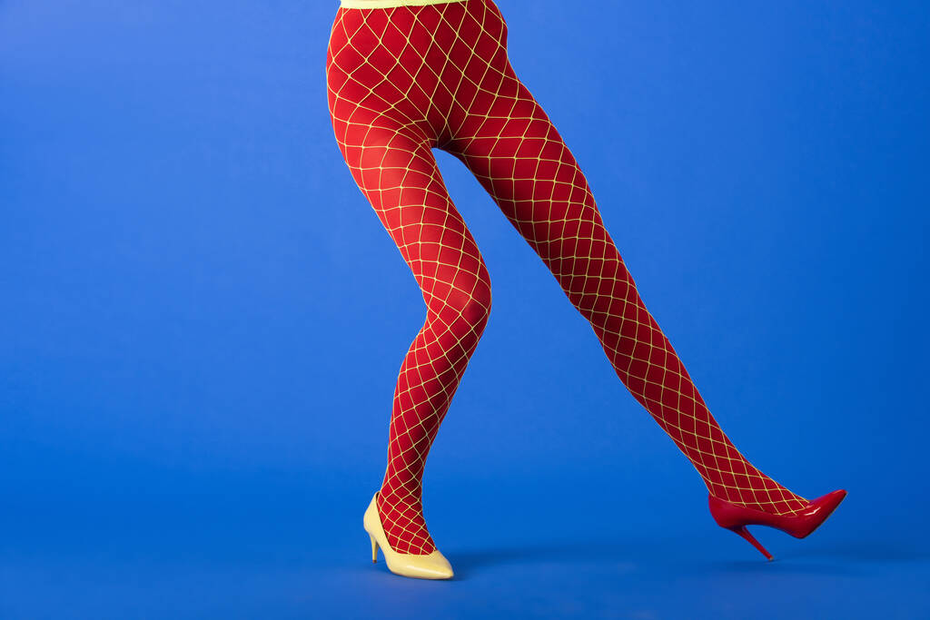 обрезанный вид модной модели в рыбных сетках колготки, желтые и красные каблуки позируя на синем
 - Фото, изображение