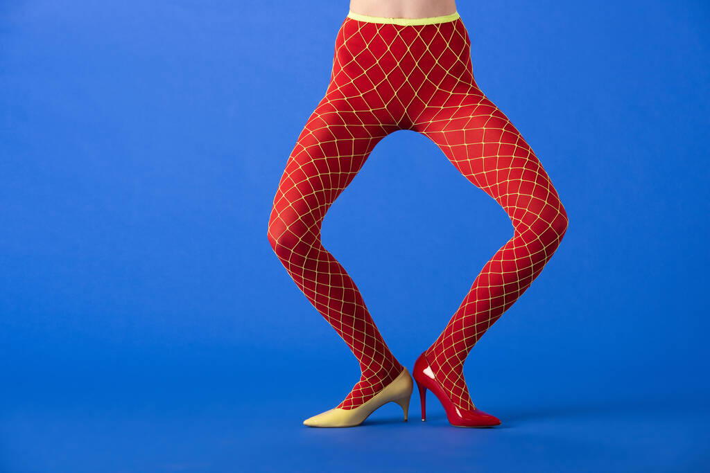 vista recortada de modelo elegante en medias de rejilla, tacones amarillos y rojos de pie en azul
 - Foto, Imagen