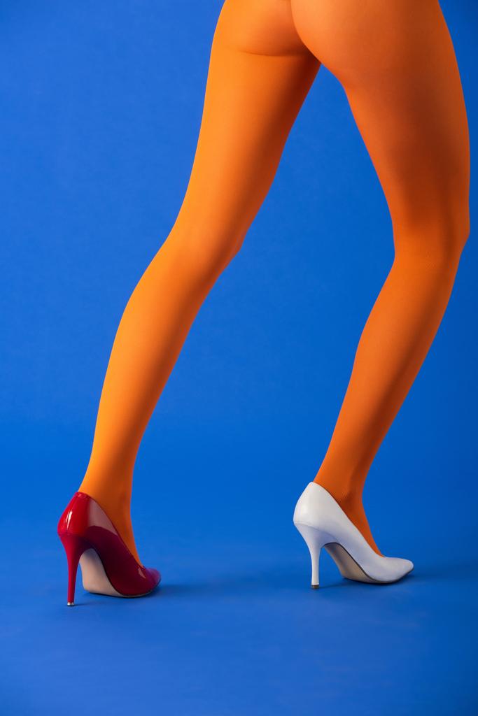 cropped άποψη της μόδας μοντέλο σε πορτοκαλί καλσόν, λευκό και κόκκινο τακούνια ποζάρουν σε μπλε - Φωτογραφία, εικόνα