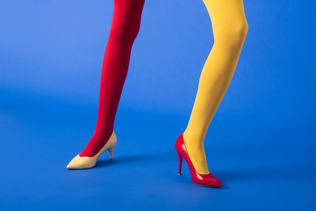 Ausgeschnittene Ansicht einer Frau in gelben und roten Strumpfhosen und Absätzen, die auf blauem Grund posiert - Foto, Bild