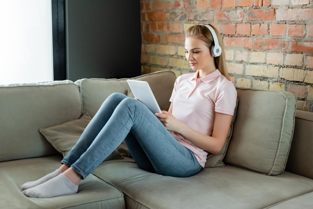 fröhliche Frau mit drahtlosen Kopfhörern mit digitalem Tablet im Wohnzimmer  - Foto, Bild