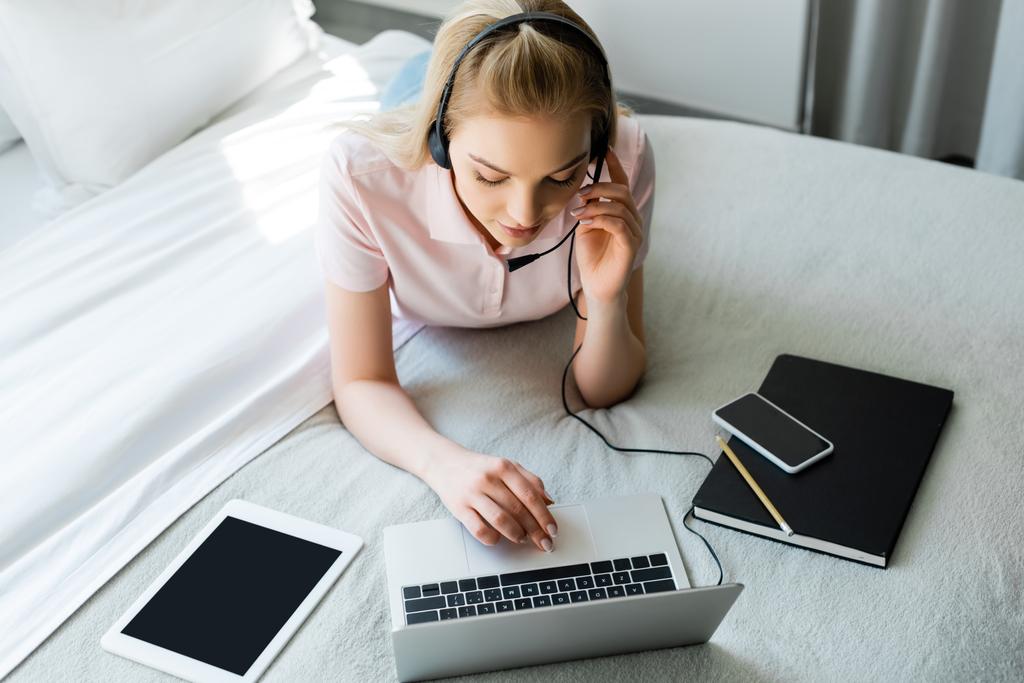 fiatal szabadúszó megható headset és a laptop közelében modulok üres képernyő az ágyon  - Fotó, kép
