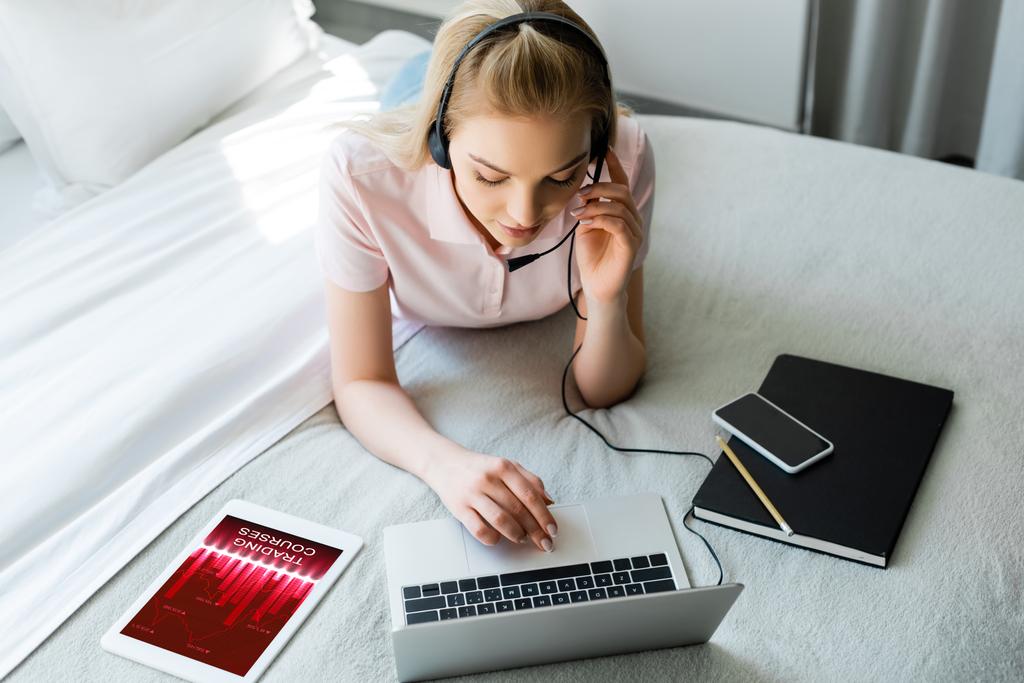 fiatal szabadúszó fülhallgató segítségével laptop közelében digitális tabletta kereskedési tanfolyamok és okostelefon üres képernyő az ágyon  - Fotó, kép
