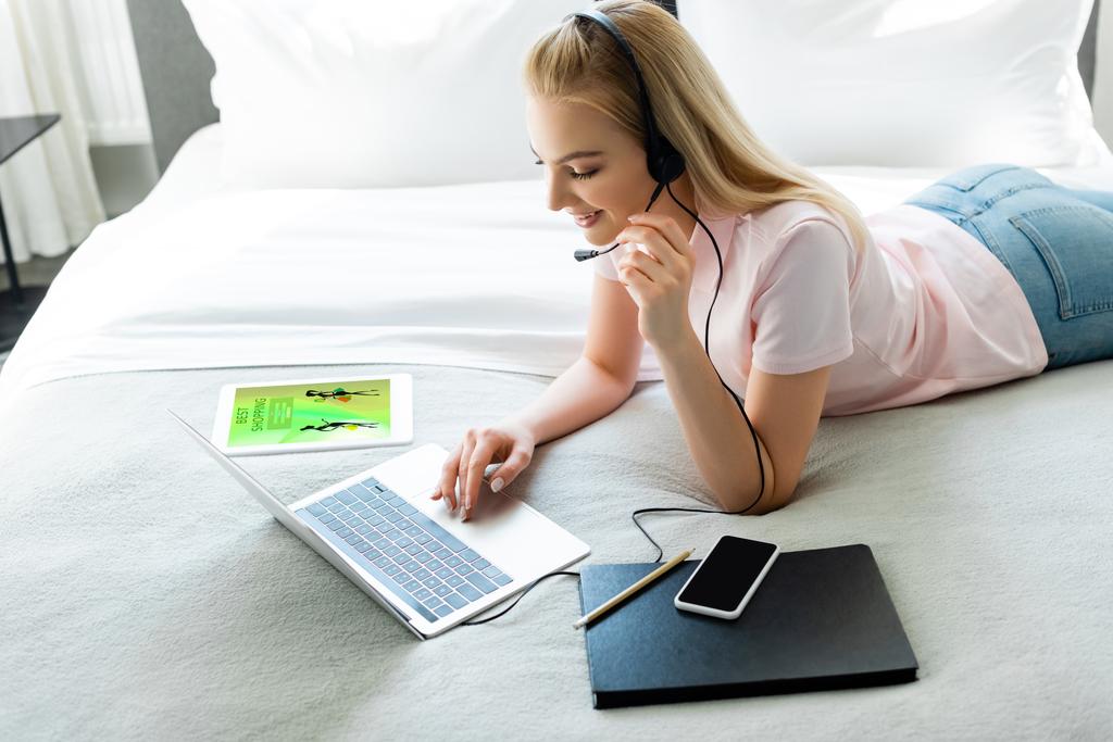 iloinen freelancer kuulokkeissa kannettavan tietokoneen lähellä digitaalista tablettia verkkokaupoissa sovellus ja älypuhelin tyhjällä näytöllä sängyssä
  - Valokuva, kuva