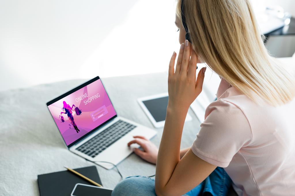 selectieve focus van freelancer aanraken headset in de buurt van laptop met online winkelen website en digitale tablet met leeg scherm op bed - Foto, afbeelding