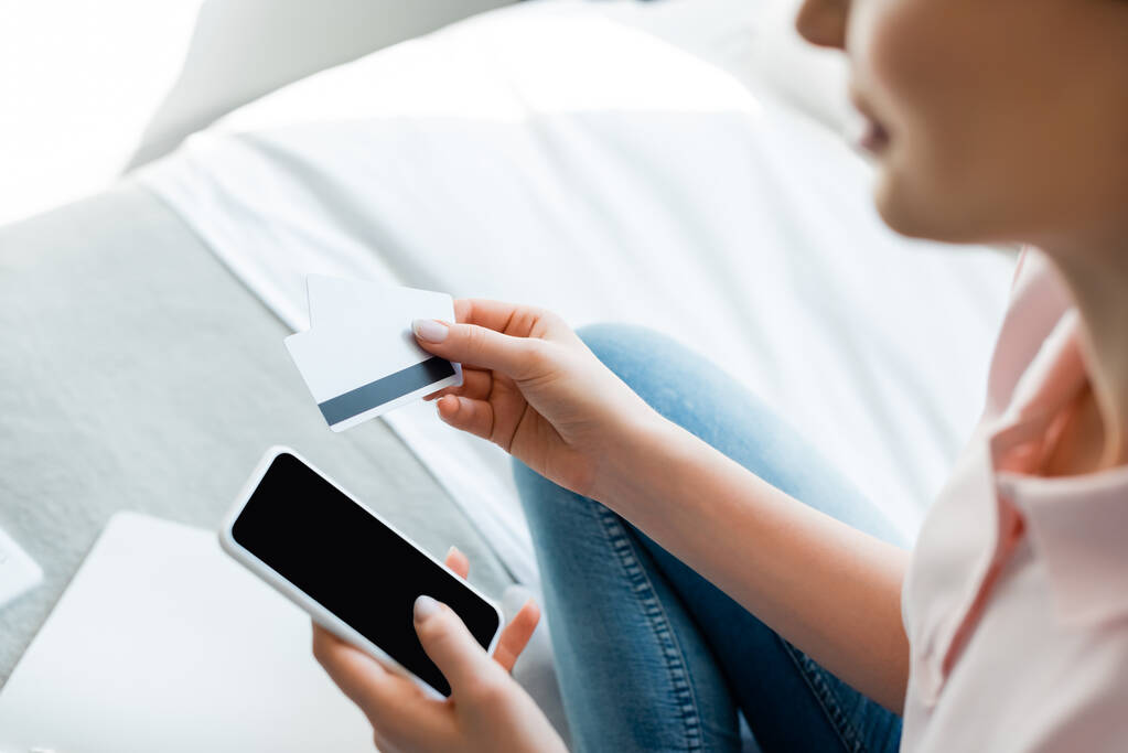επιλεκτική εστίαση της γυναίκας κρατώντας smartphone με λευκή οθόνη και πιστωτικές κάρτες στην κρεβατοκάμαρα  - Φωτογραφία, εικόνα