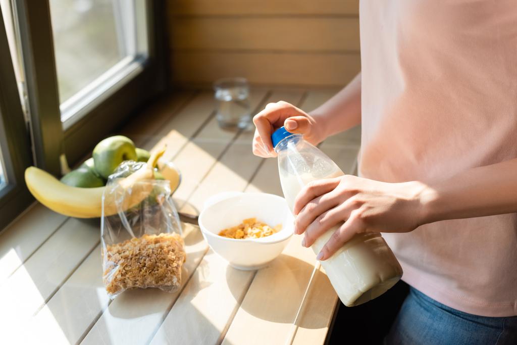 abgeschnittene Ansicht einer Frau, die eine Flasche mit Milch in der Nähe von Cornflakes in einer Schüssel und Früchten hält  - Foto, Bild