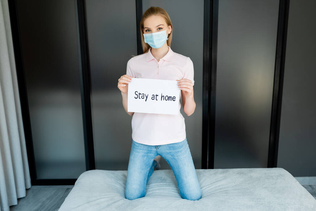 девушка в медицинской маске держит бумагу с оставшимися дома надписями
  - Фото, изображение