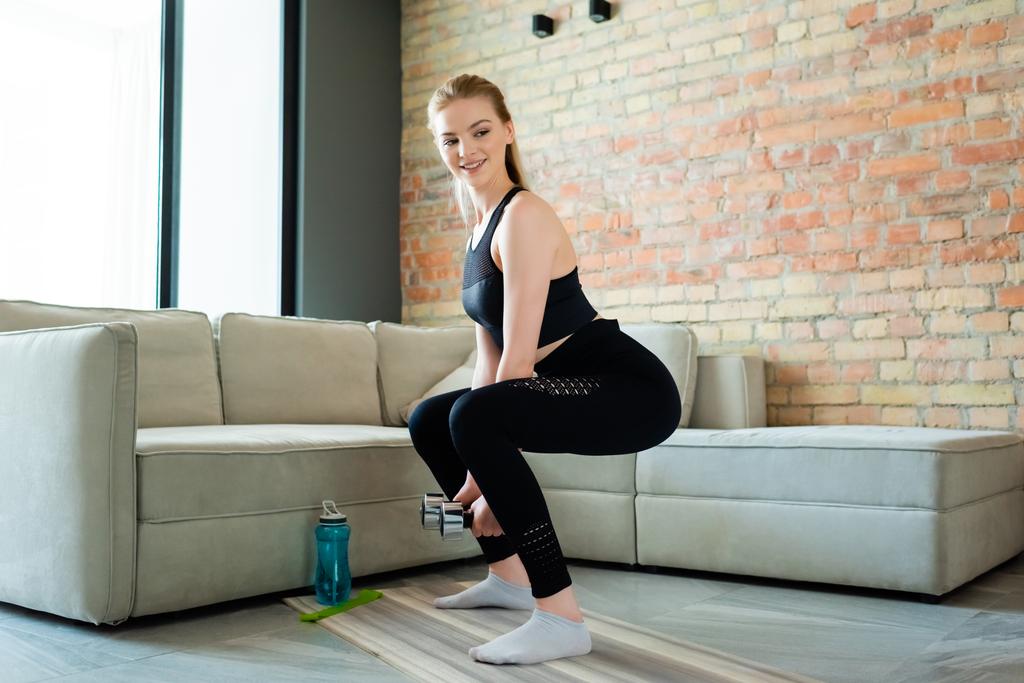 šťastný sportovkyně dělá squat cvičení s činky v blízkosti sportovní láhev v obývacím pokoji  - Fotografie, Obrázek