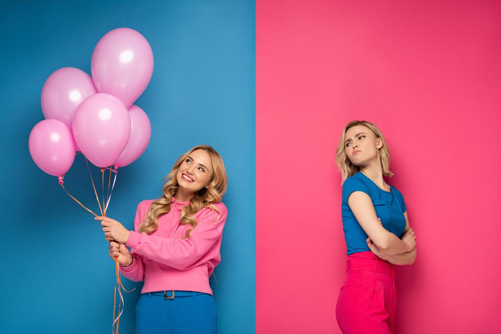 Beleidigtes blondes Mädchen blickt fröhliche Schwester mit Luftballons auf blauem und rosa Hintergrund an - Foto, Bild