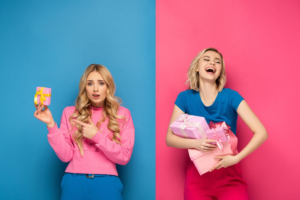 Hämmentynyt blondi nainen osoittaa lahjapakkaukseen lähellä positiivista siskoa, jolla on lahjoja sinisellä ja vaaleanpunaisella taustalla
 - Valokuva, kuva